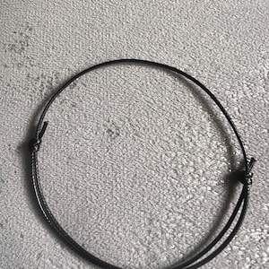 Puede incluir: Una pulsera negra ajustable hecha de un cordón fino y redondo. La pulsera está anudada en ambos extremos para su ajuste y cierre. El cordón parece estar hecho de un material liso, posiblemente encerado. La pulsera se muestra sobre una superficie texturizada de color gris claro.