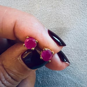 Puede incluir: Un par de pendientes de oro con tachuelas, cada uno con una piedra preciosa roja redonda y vibrante. Los pendientes se muestran sobre un fondo gris neutro, resaltando su color y diseño. El engaste de oro complementa las piedras tipo rubí.