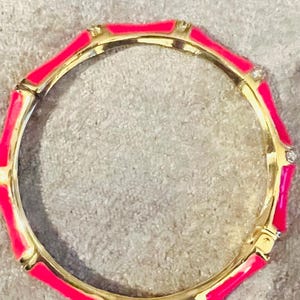May include: A gold bangle bracelet with a bright pink enamel finish. The bracelet has a geometric design with a rounded shape.