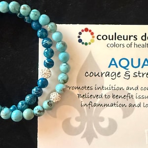 May include: Two aqua-colored beaded bracelets with silver accents. The bracelets are next to a card with the text "couleurs de santé colors of health" and "AQUA courage & strength". The card also says "TURQUOISE".
