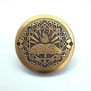 Op de afbeelding: Een gouden metalen pin met een ingewikkeld ontwerp met een boek met vlammen en een geometrisch patroon. De pin is rond en heeft een reliëfdesign.