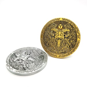 Puede incluir: Dos monedas redondas de metal con un diseño en blanco y negro de una cabeza de carnero con cuernos. La moneda de la izquierda es de plata y la moneda de la derecha es de oro. El texto "AUDERE EST-VINCERE" está inscrito en la moneda de oro.