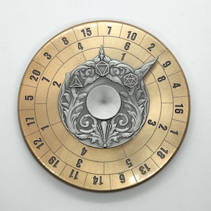 Puede incluir: Un spinner de metal de color dorado con un diseño circular. El spinner tiene un disco central de color plateado con un diseño decorativo. El anillo exterior del spinner tiene los números del 1 al 20 impresos alrededor.