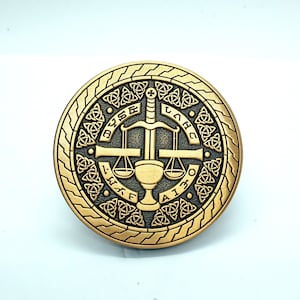 May include: A gold-colored metal coin with a Celtic knot border and a design of a sword and scales of justice. The text "DYSE LAWB AITHF" is inscribed around the design.