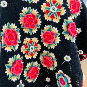 Op de afbeelding: Een zwarte gehaakte deken met kleurrijke bloemmotieven. De deken toont grote, kleurrijke bloemen in rood, roze, geel en blauw, met groene en witte accenten. De randen van de deken zijn afgewerkt met een rode rand.