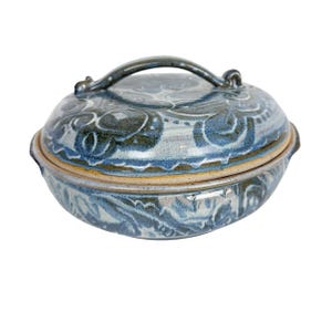 May include: A round ceramic casserole dish with a matching lid featuring a handle. The piece has a mottled blue and grey glaze with a decorative pattern of swirling shapes and lines. The base of the dish and the lid rim show a natural tan clay color.