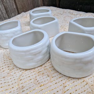 May include: A set of six white ceramic napkin rings, each featuring a smooth, glossy finish and a textured, wavy exterior surface. The rings are arranged in a cluster, showcasing their rounded, open-centre design and consistent off-white colour.