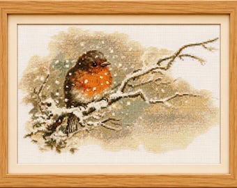 Puede incluir: Un bordado en punto de cruz enmarcado que representa un petirrojo con el pecho de color naranja brillante posado sobre una rama cubierta de nieve. La escena muestra copos de nieve blancos cayendo sobre un fondo suave beige y verde, en un marco de madera clara.
