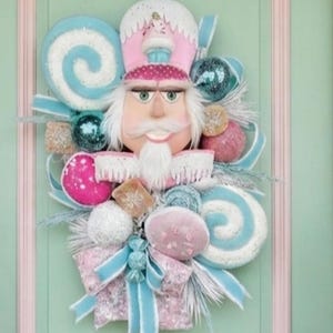 May include: A pastel-themed holiday display featuring two large pink and light blue nutcracker figures standing on either side of a door, with a matching nutcracker-shaped wreath centered on the door, all surrounded by frosted garland with candy ornaments.