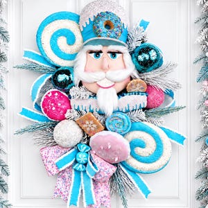 May include: A decorative nutcracker wreath featuring a central nutcracker figure adorned with a donut hat, surrounded by blue and pink candy-themed ornaments, glittery lollipops, frosted pine branches, and a patterned bow with light blue ribbons.