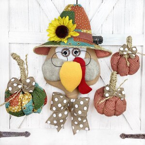 May include: A patchwork turkey door hanger with a brown, yellow, and red face. The turkey is wearing a brown, green, and orange patchwork hat with a yellow sunflower. The turkey is holding a brown and white polka dot bow. There are three red and white gingham pumpkins hanging from the turkey.
