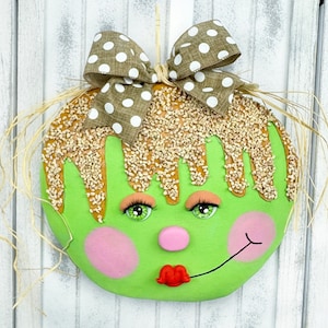 May include: A green and brown felt door hanger shaped like a face with a brown bow, brown hair, green eyes, a pink nose, and a red mouth. The face is decorated with brown sprinkles.