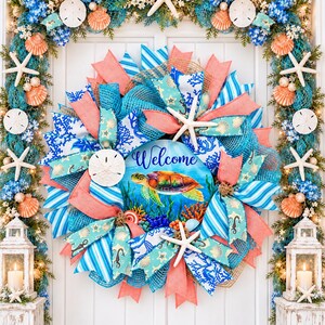 May include: A decorative wreath featuring a central sign with a sea turtle illustration and the word Welcome, surrounded by blue and coral mesh ribbons, white starfish, and sand dollars, framed by matching garlands and two white lanterns.