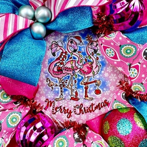K&ouml;nnte beinhalten: Ein rosa-blauer Weihnachtskranz mit einem festlichen Design, das drei rosa Flamingos zeigt, die Weihnachtsm&uuml;tzen tragen und Zuckerstangen halten. Der Kranz ist mit rosa und blauen Ornamenten, Glitzer und einem Schild verziert, auf dem "Frohe Weihnachten" steht.