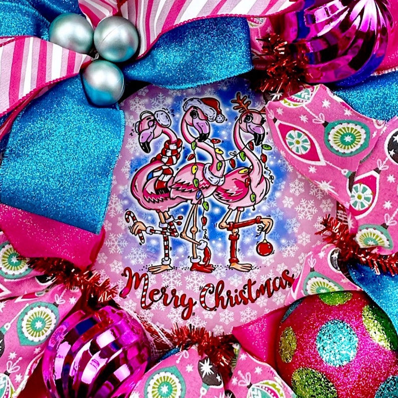 K&ouml;nnte beinhalten: Ein rosa-blauer Weihnachtskranz mit einem festlichen Design, das drei rosa Flamingos zeigt, die Weihnachtsm&uuml;tzen tragen und Zuckerstangen halten. Der Kranz ist mit rosa und blauen Ornamenten, Glitzer und einem Schild verziert, auf dem "Frohe Weihnachten" steht.