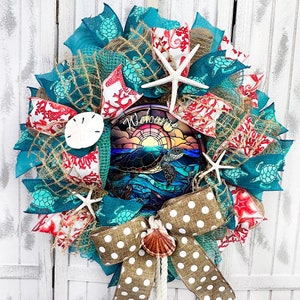 May include: A decorative wreath with a coastal theme. It features turquoise and red ribbons, starfish, a sand dollar, and a bow. The center has a stained-glass-style image of a sea turtle and the word "Welcome".