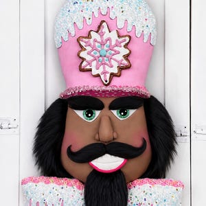 May include: A decorative nutcracker wall hanging featuring a pink hat topped with white glittery frosting and sprinkles. The nutcracker has dark skin, green eyes, a black mustache and beard, and a white snowflake emblem on the front of the hat.