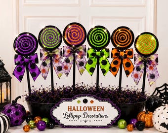 Peut inclure: Six sucettes décoratives sur bâtonnets, chacune avec un motif en spirale violet, vert ou orange, agrémentées de nœuds à motifs. Un panneau central indique HALLOWEEN Lollipop Decorations, entouré de petites sphères scintillantes et citrouilles.