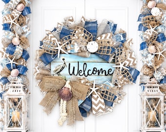May include: A coastal-themed wreath with a "Welcome" sign, starfish, and seashells. The wreath features blue and tan ribbons, burlap, and decorative netting. A matching garland and lanterns flank the door, creating a welcoming entryway.