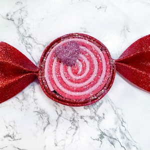 May include: A red and pink candy-shaped ornament with a swirl design and a small pink heart on top. The ornament is made of felt and has a glittery finish.