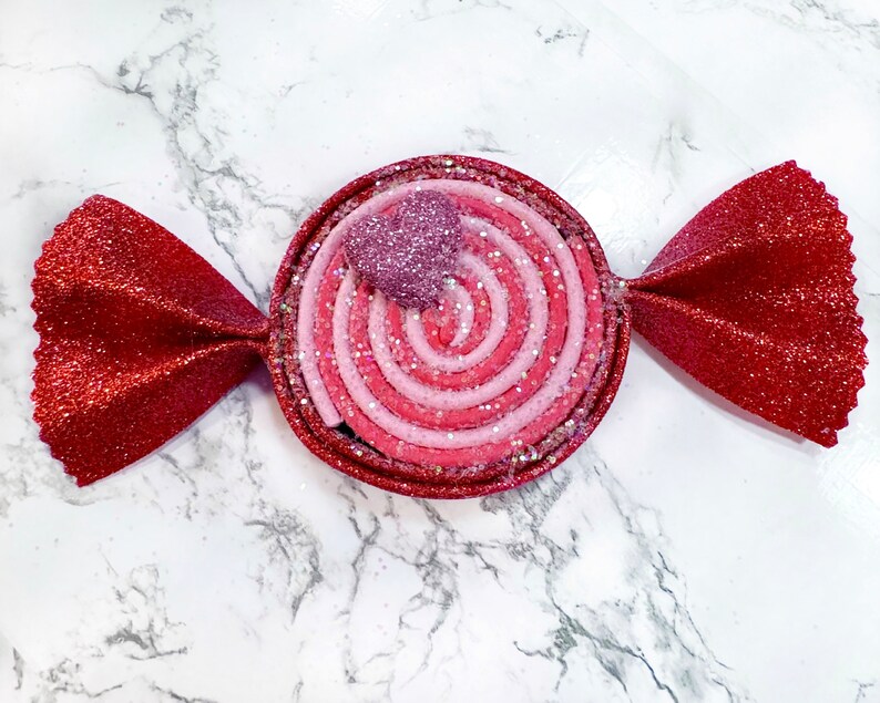 May include: A red and pink candy-shaped ornament with a swirl design and a small pink heart on top. The ornament is made of felt and has a glittery finish.