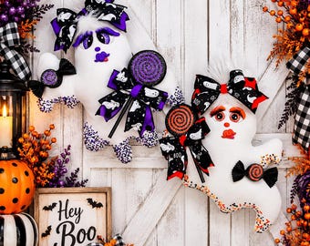 Peut inclure: Deux décorations fantômes en peluche avec des corps en tissu blanc, des accents violets et orange, et des nœuds à motifs. Chaque fantôme tient une sucette scintillante et présente des traits peints. Le fantôme de gauche a des détails violets, celui de droite des accents orange et un panneau indiquant Hey Boo.
