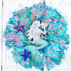 May include: A decorative mermaid wreath with a white mermaid figure holding a seashell. The wreath features turquoise, blue, and pink ribbons, starfish, and coral accents. The wreath is approximately 20 inches in diameter.