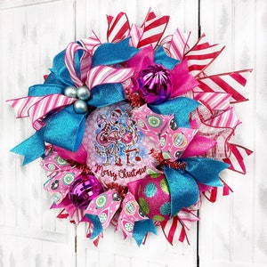 K&ouml;nnte beinhalten: Ein rosa-blauer Weihnachtskranz mit einem Flamingo-Motiv und dem Text "Merry Christmas". Der Kranz ist mit rosa-wei&szlig; gestreiftem Band, blauem Glitzerband und rosa-wei&szlig;en Kugeln verziert.