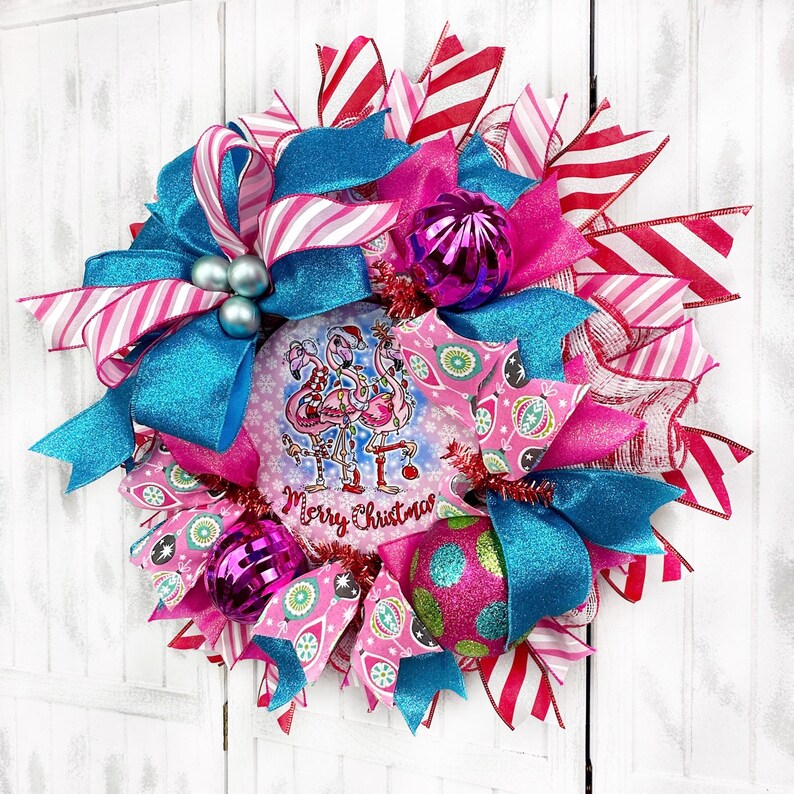 K&ouml;nnte beinhalten: Ein rosa-blauer Weihnachtskranz mit einem Flamingo-Motiv und dem Text "Merry Christmas". Der Kranz ist mit rosa-wei&szlig; gestreiftem Band, blauem Glitzerband und rosa-wei&szlig;en Kugeln verziert.