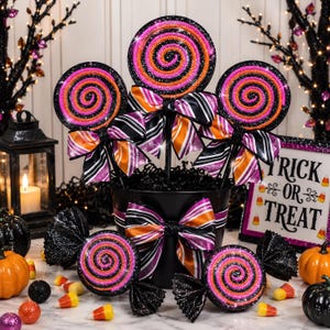 May include: A black bucket filled with three large lollipop-shaped decorations featuring orange, pink, and black glittery swirls, each accented with a striped bow. Two additional candy-shaped decorations sit in front, with a sign reading Trick or Treat.