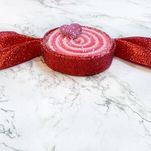 May include: A red and pink candy cane shaped ornament with a glitter finish. The candy cane is topped with a pink heart.