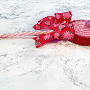 May include: A red and pink glittery swirl lollipop with a red ribbon bow with white polka dots and pink hearts. The lollipop is on a white marble surface.