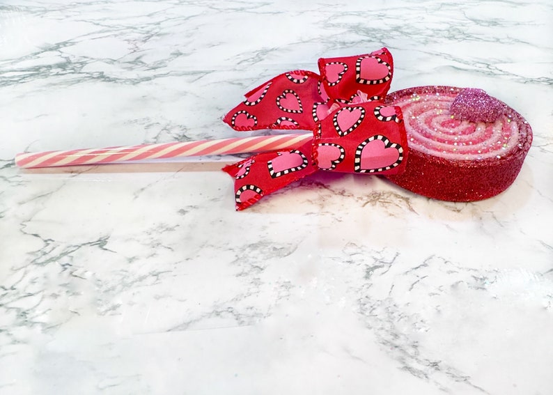 May include: A red and pink glittery swirl lollipop with a red ribbon bow with white polka dots and pink hearts. The lollipop is on a white marble surface.