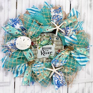 May include: A blue and white nautical themed wreath with a sign that says "Welcome to the River". The wreath is decorated with starfish, seashells, and fishing nets.