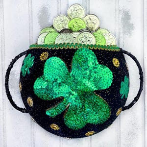 May include: A black pot with a green shamrock design and gold sequins. The pot is filled with gold coins with shamrock designs.