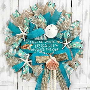 May include: A turquoise, brown, and white wreath with a sign that reads "Meet me where the sand touches the sea". The wreath is decorated with seashells, starfish, and burlap ribbon.