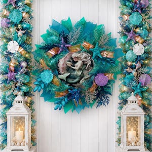 May include: A mermaid-themed wreath with a central mermaid figurine, surrounded by teal and turquoise mesh, seashells, and starfish. The wreath is flanked by matching garland with lights and white lanterns with candles, creating a coastal theme.
