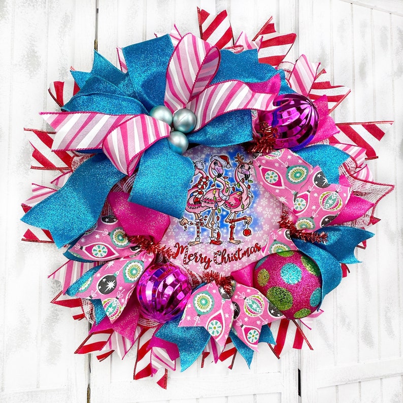 K&ouml;nnte beinhalten: Ein rosa-blauer Weihnachtskranz mit einem Flamingo-Motiv und dem Text "Merry Christmas". Der Kranz ist mit rosa-blauen Schleifen, Glitzer und Kugeln verziert.