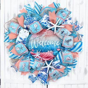 May include: A welcome wreath with a blue and white ocean-themed design. The wreath is decorated with seashells, starfish, coral, and a welcome sign. The wreath is made with burlap, mesh, and ribbon.