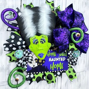 May include: A Halloween wreath featuring a green Frankenstein-themed character with white hair, a purple spiderweb bow, and a sign that reads "Home Sweet Haunted Home".