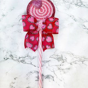 May include: A pink and red glitter swirl lollipop with a pink heart on top, attached to a pink and white striped stick with a red bow with white polka dots and pink hearts.