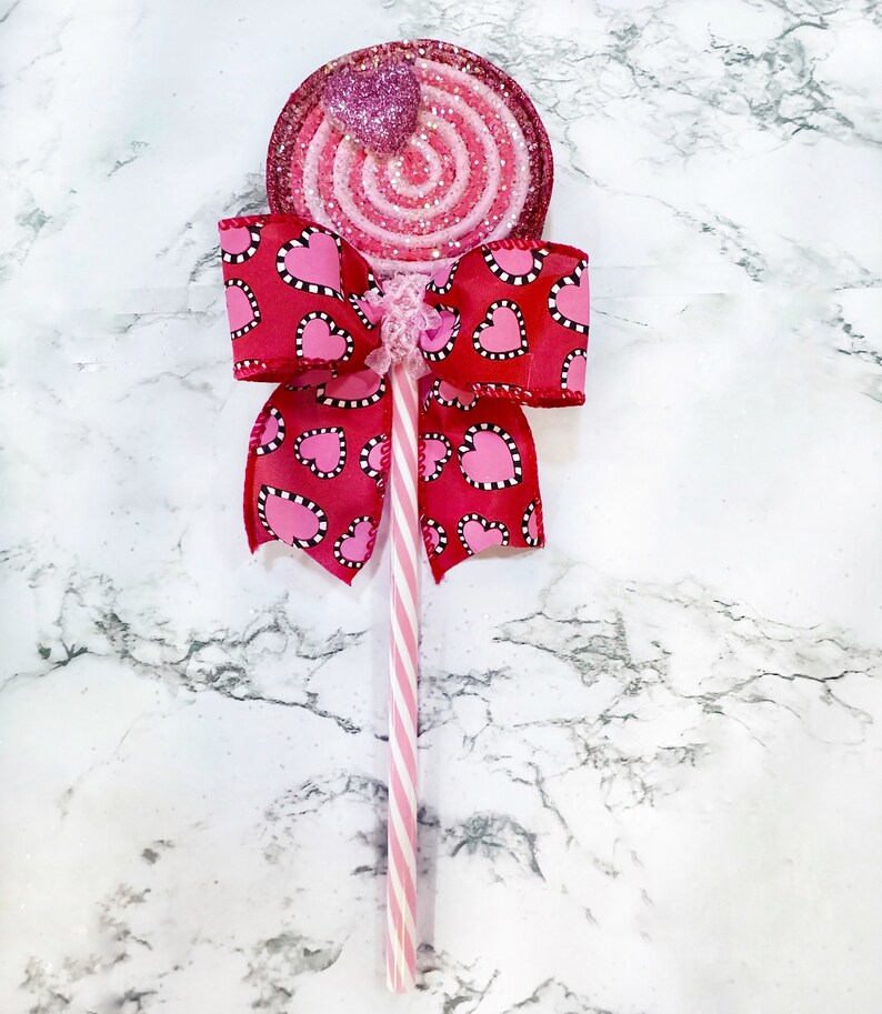 May include: A pink and red glitter swirl lollipop with a pink heart on top, attached to a pink and white striped stick with a red bow with white polka dots and pink hearts.