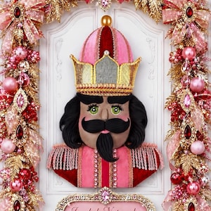 May include: A nutcracker wreath attachment featuring a pink and red velvet uniform with gold trim, a gold crown, and black hair. The figure is surrounded by a decorative wreath of pink and red ornaments, gold leaves, and ribbons with a sign reading Jeweled Nutcracker Wreath Attachment.
