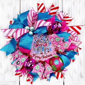 K&ouml;nnte beinhalten: Ein rosa-blauer Weihnachtskranz mit einem Flamingo-Motiv und dem Text "Merry Christmas". Der Kranz ist mit rosa, wei&szlig;em und rotem Band verziert und hat verschiedene Ornamente.