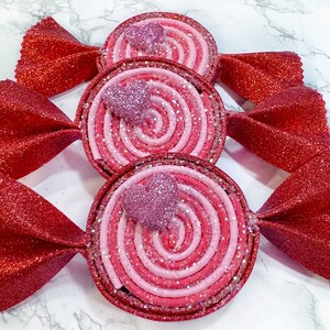 May include: Three red and pink candy-shaped ornaments with glitter. Each ornament has a pink heart in the center and a swirl design.
