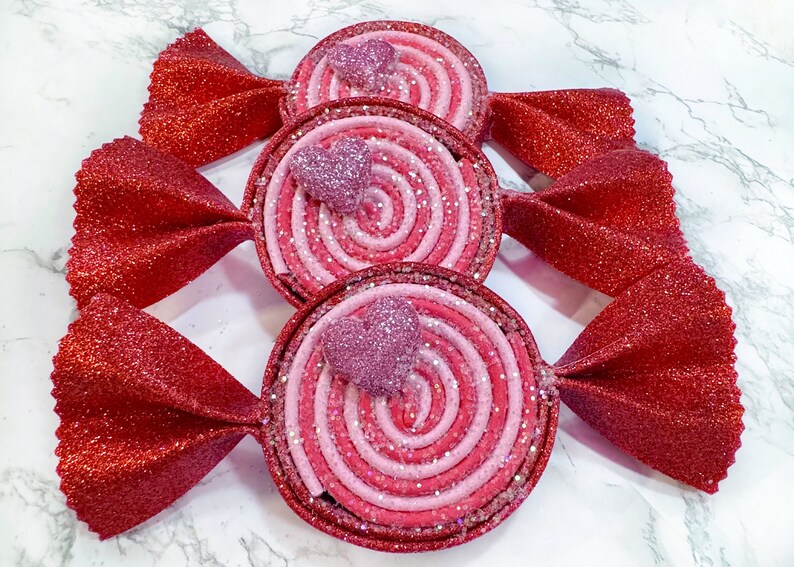May include: Three red and pink candy-shaped ornaments with glitter. Each ornament has a pink heart in the center and a swirl design.