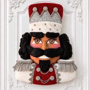 May include: A decorative nutcracker wall hanging featuring a red and white uniform with silver fringe epaulets. The figure has a black yarn mustache and beard, a crown with silver glitter and rhinestones, and a white faux fur hat trim.