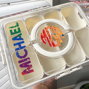 May include: A clear plastic lunch box with multiple compartments and a white lid. The lid features a white circle with a colorful monogram and the name "MICHAEL" in rainbow-colored letters. The lunch box is designed for food storage.