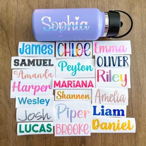 May include: A light purple water bottle with a black lid and the name Sophia in iridescent lettering, surrounded by numerous rectangular white vinyl decals featuring various names in different colors, fonts, and finishes like glitter and metallic.