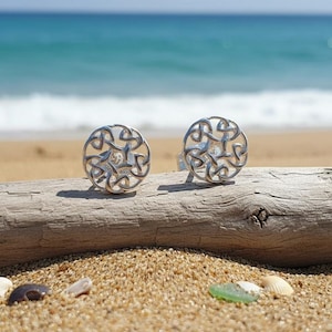 Può includere: Due orecchini a lobo in argento con un design a nodo celtico traforato, ciascuno con una pietra trasparente al centro. Gli orecchini sono esposti su un pezzo di legno trasportato dal mare, con una spiaggia sabbiosa e l'oceano sullo sfondo.